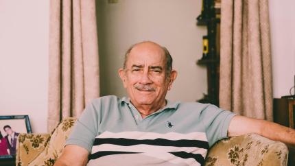 An older man sits on a chair and smiles at a camera