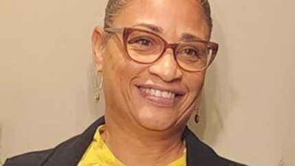 Idella Winfield headshot, with Idella wearing red glasses, earrings, a black jacket, and a bright yellow blouse