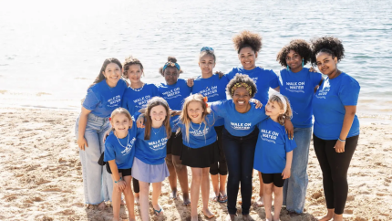 A group of girls stands on a beach, smiling. They all are wearing blue shirts that say "walk on water"
