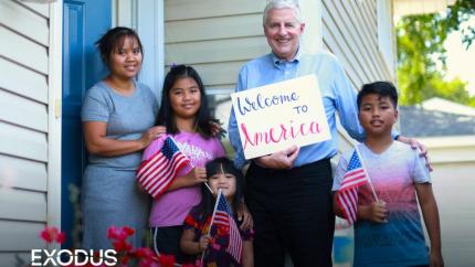 A refugee family welcomed by a sponsor as they hold American flags and a sign of welcome