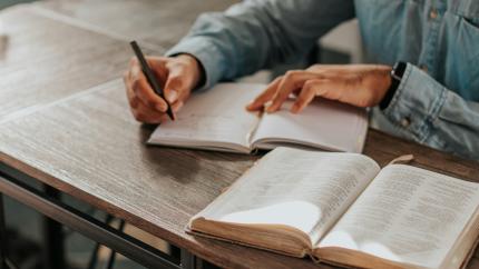 An individual sits at a wooden desk, reading a Bible. Courtesy of Fa Barboza on Unsplash.