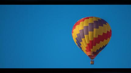 a red, yellow, and blue checkered hot air balloon lifts off into the blue sky