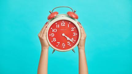 Two hands hold up an analogue alarm clock in front of a teal background