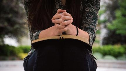 Individual sits on a bench, reading the biblical text with her hands crossed in prayer.