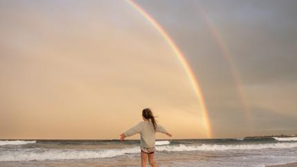 A young women raises her arms while running into the surf under a rainbow on the beach.
