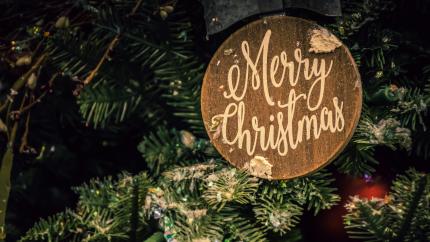 A plaque that reads "Merry Christmas" sits on an evergreen tree. Courtesy of Brett Sayles on Pexels.