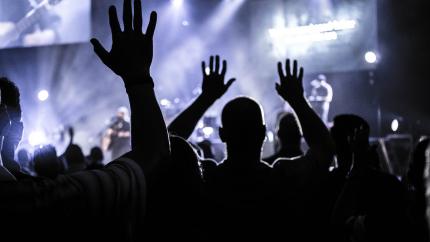 A group of worshippers raises their hands in a large concert setting.