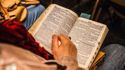 An open Bible rests on someone's lap; their hand holds open the pages.