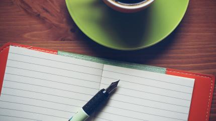 A notebook, pen, and coffee cup sit on a wood surface.