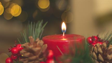 Red advent candle with a yellow flame burns surrounded by pinecones and winter berries