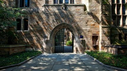 A gate to a large university campus is shown, with a large, white stone building behind it. Courtesy of Pixaby.
