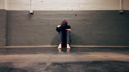 a young adult sits against a wall. His arms are crossed on top of his knees and his head is down.