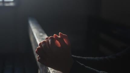 An individual stands with their hands folded on the front of a church pew. Courtesy of Pixaby.