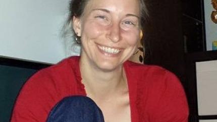 Anita sits, smiling, wearing a red shirt and jeans