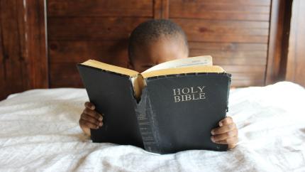 small child reading large Bible in bed
