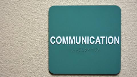 A plaque with the word "Communication" in both English text and Braille text hands on a white wall. Courtesy of iStock.