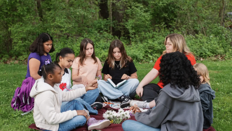 Image of a group of girls doing bible studies outside on a green lawn with a green wood in the background. Courtesy of GEMS.