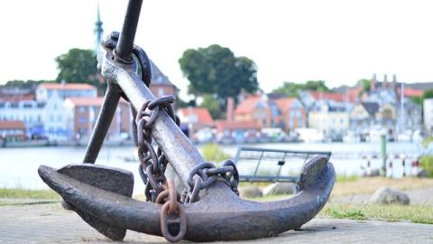 An anchor sits on a platform outside a seaside town. Courtesy of Pixaby.