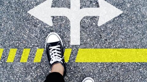 Standing at a crossroads, a person with black shoes looks at the pavement and sees a three-directional arrow