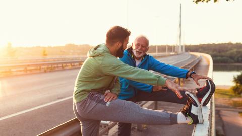 In the pedestrian lane of a bridge, an elderly man and a young man, both in athletic gear, stretch their legs