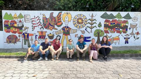 A group of university students sit on a curb in front of a mosaic in El Salvador