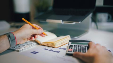 A man holds a pen and a calulcator as he works on a bookkeeping project. Courtesy of Unsplash.