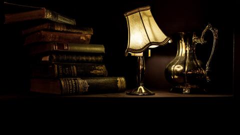 A stack of books, an old lit lamp, and an antique pitcher sit on a shelf