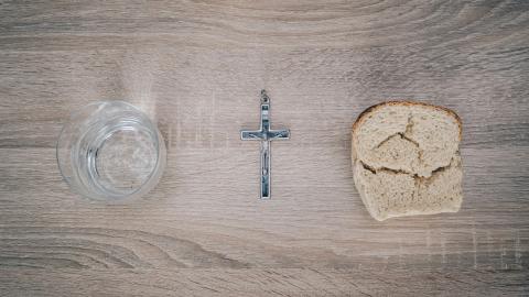 A photo of a glass, a cross, and a piece of bread on a table.