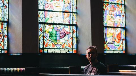 a young person sits in a pew in front of stained glass windows