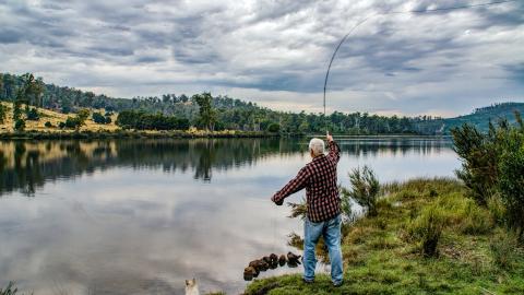 A man stands on the side of a lake and throws his fishing road over the lake