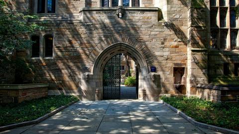 A gate to a large university campus is shown, with a large, white stone building behind it. Courtesy of Pixaby.