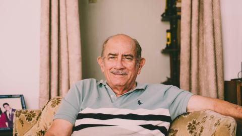 An older man sits on a chair and smiles at a camera