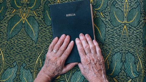 On a table cloth, old hands rest on a black book with the word "Biblia" in gold lettering