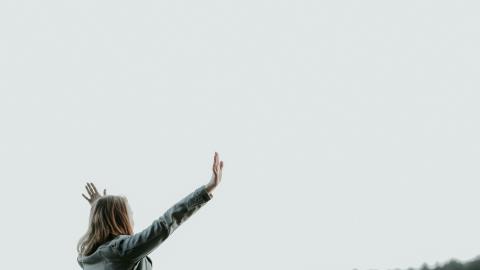 A woman holds her hands up in praise, with a white sky in the background. Courtesy of Unsplash