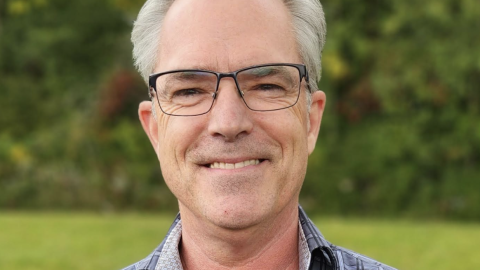 Ron deVris headshot, wearing a blue plaid shirt an eyeglasses on a green background